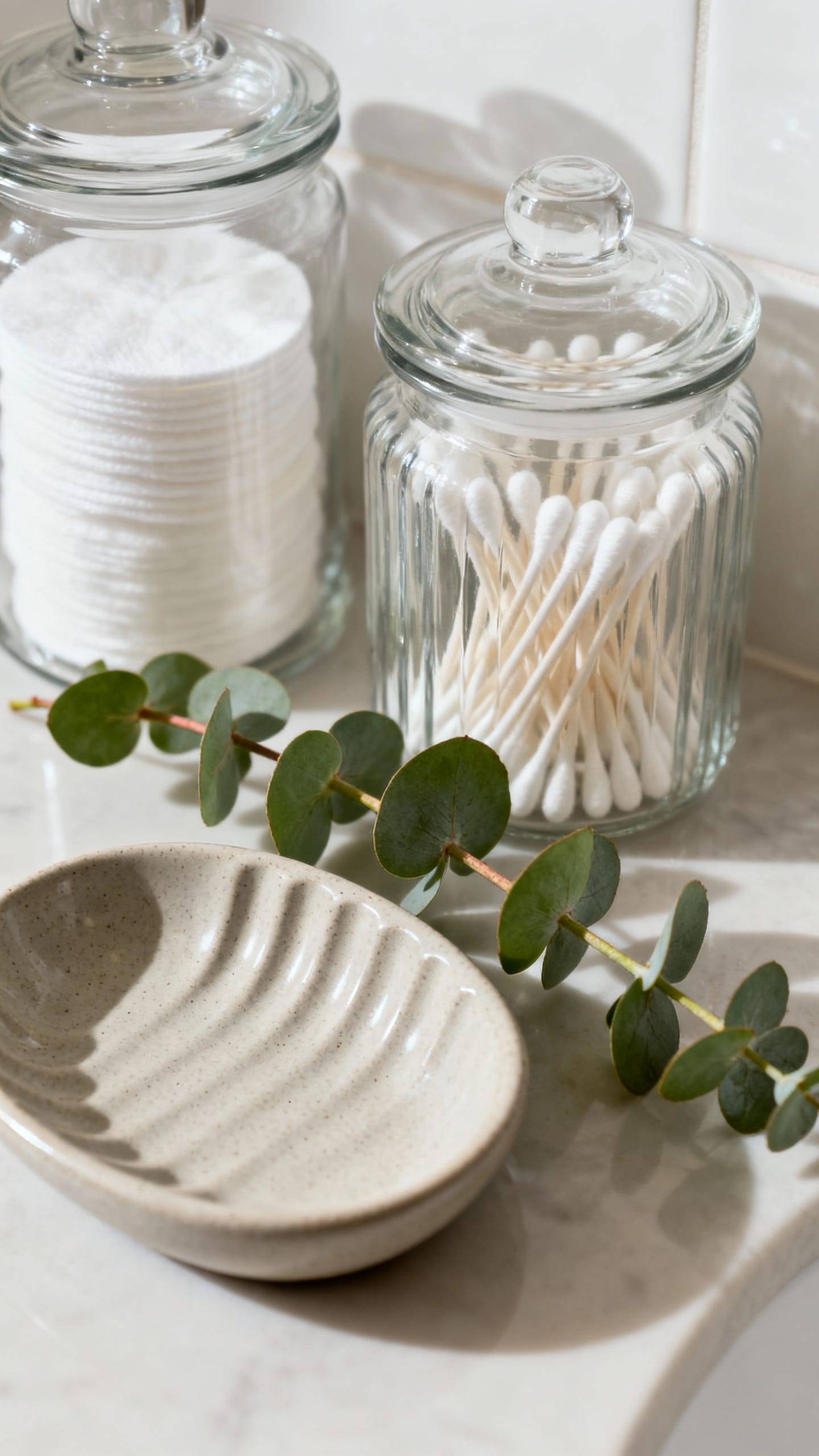 Bathroom counter closeup: glass jars with cotton rounds and swabs, ridged ceramic soap dish, eucalyp